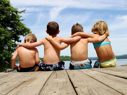 4 amigos, son pequeños, que enlazan sus brazos entre sí para estar unidos, sentados en un "muelle" mirando el horizonte.